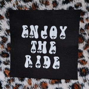 Peut inclure: Patch en tissu noir avec du texte blanc qui dit "Enjoy the Ride" dans une police rétro. Les lettres sont décorées de petites illustrations de champignons.