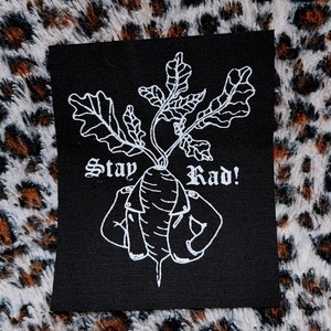 以下が含まれることがあります： 白い刺繍デザインの葉と「Stay Rad！」の文字が入った黒い正方形のパッチ。 大根は腕を曲げて様式化されています。 パッチはヒョウ柄の背景にあります。