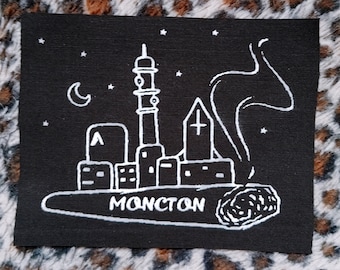 Könnte beinhalten: Ein schwarzes Leinwandbild mit weißer Farbe, das eine Skyline mit einem hohen Gebäude, einem Halbmond und Sternen darstellt. Die Stadt ist unten mit "MONCTON" beschriftet.