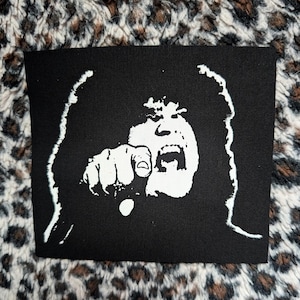 Könnte beinhalten: Ein rechteckiger schwarzer Stoffaufnäher mit einer kontrastreichen weißen Siebdruckgrafik einer Person mit langen Haaren, offenem Mund und einer nach vorne ausgestreckten Hand mit zeigendem Finger. Das Design wirkt wie eine Schablone.