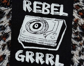 Puede incluir: Parche termoadhesivo blanco y negro con un tocadiscos y el texto "REBEL GRRRL" en estilo manuscrito.