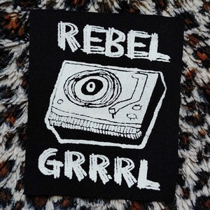 May include: Black and white iron-on patch featuring a record player and the text "REBEL GRRRL" in a handwritten style.