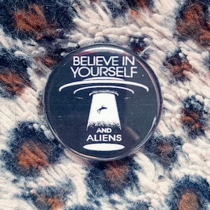 May include: A black and white button with the text "Believe in Yourself. And Aliens" and a black and white illustration of a UFO with a person falling from it.