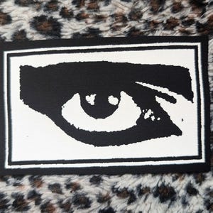 May include: A rectangular fabric patch featuring a high-contrast black and white graphic design. The central image depicts a stylized, singular human eye with a bold black iris and pupil set against a white background, framed by a thick black border.
