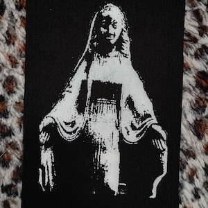 May include: A black and white iron-on patch featuring a stylized image of a woman in a long robe with her arms outstretched.