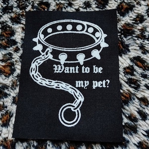 May include: A black fabric patch with white text and a white image of a spiked collar with a chain attached. The text reads "Want to be my pet?"