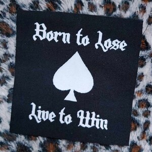 Puede incluir: Parche de tela negro con texto blanco que dice "Born to Lose Live to Win" y un símbolo de picas blanco en el centro.