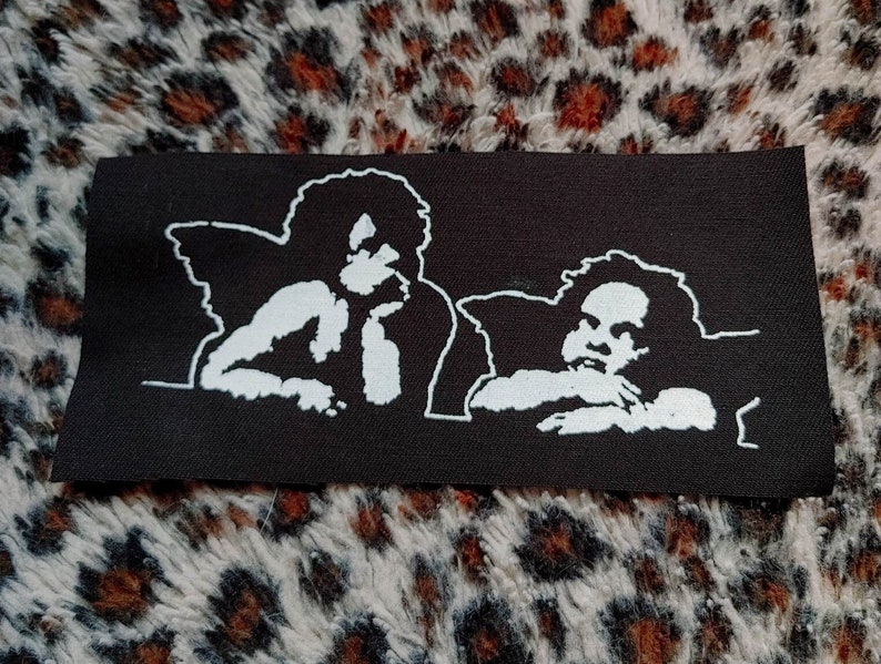 May include: Black and white embroidered patch featuring two angels with wings, one looking to the side and the other looking straight ahead.