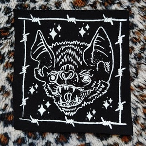 May include: A black and white embroidered patch of a bat's head with sharp teeth and glowing eyes. The bat is surrounded by a barbed wire frame.