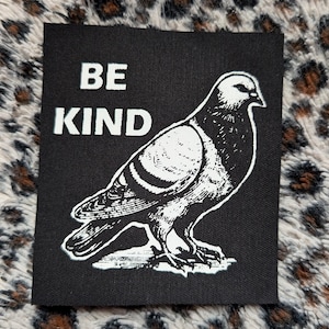 以下が含まれることがあります： 白い文字で「BE KIND」と書かれた黒い正方形のパッチ。その下に、白いハトの精巧なイラストが描かれています。パッチはヒョウ柄の背景に配置されています。
