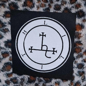 May include: A black square patch with a white circular emblem. The emblem has a central symbol with cross-like features and the letters "L", "I", and "H" around the edge. Suitable for application on clothing or accessories.