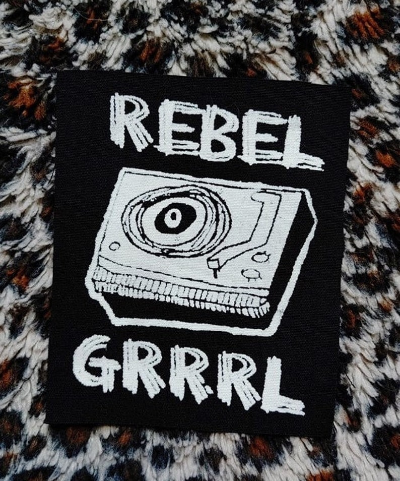 May include: Black and white iron-on patch with a record player and the text "REBEL GRRRL" in a handwritten style.
