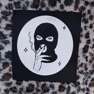 May include: Black and white patch with a circle design. Inside the circle is a person wearing a black ski mask and holding a cigarette. There are three white stars around the circle.