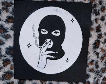 Könnte beinhalten: Schwarz-weißes Patch mit einem Kreisdesign. Im Kreis befindet sich eine Person mit einer schwarzen Skimaske, die eine Zigarette hält. Um den Kreis herum befinden sich drei weiße Sterne.