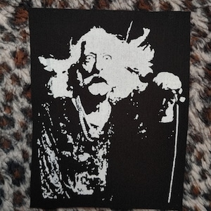 May include: Black and white iron-on patch featuring a silhouette of a man with long hair and a beard, holding a microphone. The patch is designed for sewing onto clothing or bags.