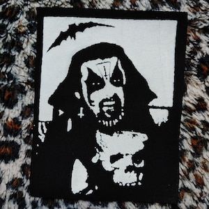 Puede incluir: Parche bordado en blanco y negro con una figura encapuchada que sostiene una calavera en la mano. La figura tiene una expresión amenazante y lleva un casco con cuernos. El parche está sobre un fondo negro.