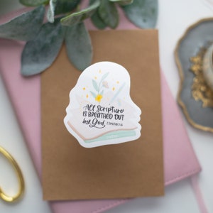 May include: A white sticker with the text "All scripture is breathed out by God" and the Bible verse 2 Timothy 3:16. The sticker features a floral design with a pencil and a stack of books. The sticker is on a brown card.