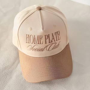 Op de afbeelding: Beige baseballpet met een bruine klep en de geborduurde tekst "HOME PLATE Social Club". De pet heeft een gebogen klep en een knoop aan de bovenkant. De hoed is gemaakt van een zacht materiaal.