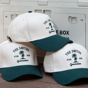May include: Cream and dark green baseball caps. Each cap displays "THE SMITHS", a palm tree graphic, and the date "07.08 2026". The word "Groomsman" is printed below. The caps are stacked on a white storage box, suitable for a wedding.