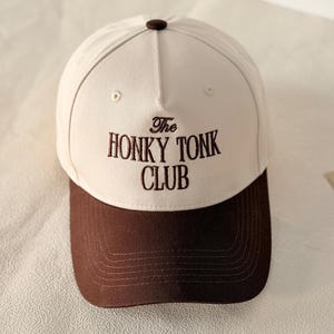 May include: A cream-coloured baseball cap with a dark brown brim and a dark brown button on top. The front of the cap features dark brown embroidered text that reads The HONKY TONK CLUB in a serif font. The cap has five panels and eyelets.