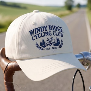 Könnte beinhalten: Eine cremeweiße Baseballkappe mit dunkelblauem Schriftzug Windy Ridge Cycling Club, einer Fahrradillustration auf einer Straße, Tannenbäumen und dem Text Est. 2023, die auf dem Lenker eines Fahrrads mit braunen Ledergriffen liegt.