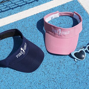 Könnte beinhalten: Zwei Sonnenblenden und herzförmige Sonnenbrillen liegen auf einer blauen Oberfläche. Eine Sonnenblende ist marineblau, die andere rosa. Beide Sonnenblenden tragen den Text "FORE GIRLS" in Weiß. Die Sonnenbrille ist hellblau.