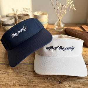 Op de afbeelding: Een marineblauwe zonneklep met de witte geborduurde tekst the party en een witte zonneklep met de donkerblauwe geborduurde tekst wife of the party. Beide kleppen hebben een gebogen rand en een gestructureerd voorpaneel met stiksels.