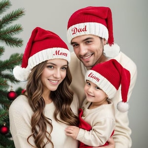 Könnte beinhalten: Eine Familie trägt passende rot-weiße Weihnachtsmützen. Auf den Mützen stehen die Wörter "Dad", "Mom" und "Liam". Die Familie lächelt vor einem Weihnachtsbaum.