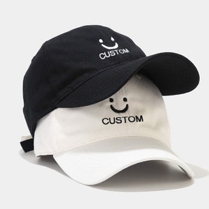 Pode incluir: Um boné de beisebol reversível em preto e branco com um rosto sorridente e a palavra "CUSTOM" bordada em ambos os lados.