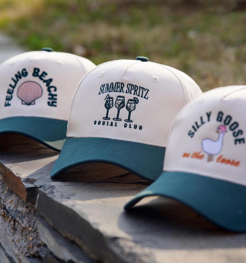May include: Three beige baseball caps with green brims. The caps have different embroidered designs: "Feeling Beachy" with a seashell, "Summer Spritz Social Club" with three cocktail glasses, and "Silly Goose on the Loose" with a goose.