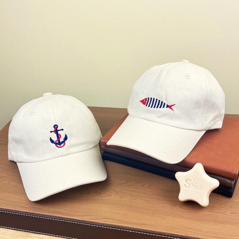 Puede incluir: Dos gorras de b&eacute;isbol beige con dise&ntilde;os bordados. Una gorra presenta un ancla azul con una cuerda roja, la otra un pez rayado rojo y azul. Ambas gorras est&aacute;n sobre una superficie de madera con una pastilla de jab&oacute;n en forma de estrella.