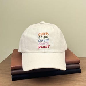 Puede incluir: Una gorra de b&eacute;isbol blanca con las palabras "Cheers", "Salud", "Cin Cin", "Sant&eacute;" y "Prost" bordadas en diferentes colores.