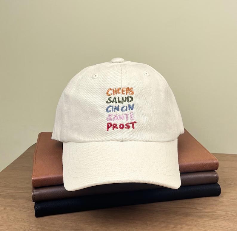 Puede incluir: Una gorra de b&eacute;isbol blanca con las palabras "Cheers", "Salud", "Cin Cin", "Sant&eacute;" y "Prost" bordadas en diferentes colores.