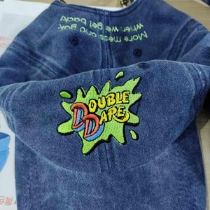 Puede incluir: Una gorra de b&eacute;isbol de mezclilla azul con un logotipo bordado en verde, amarillo y rojo que dice "Double Dare". La parte posterior de la gorra tiene texto blanco que dice "M&aacute;s desorden y m&aacute;s diversi&oacute;n, cuando volvamos"