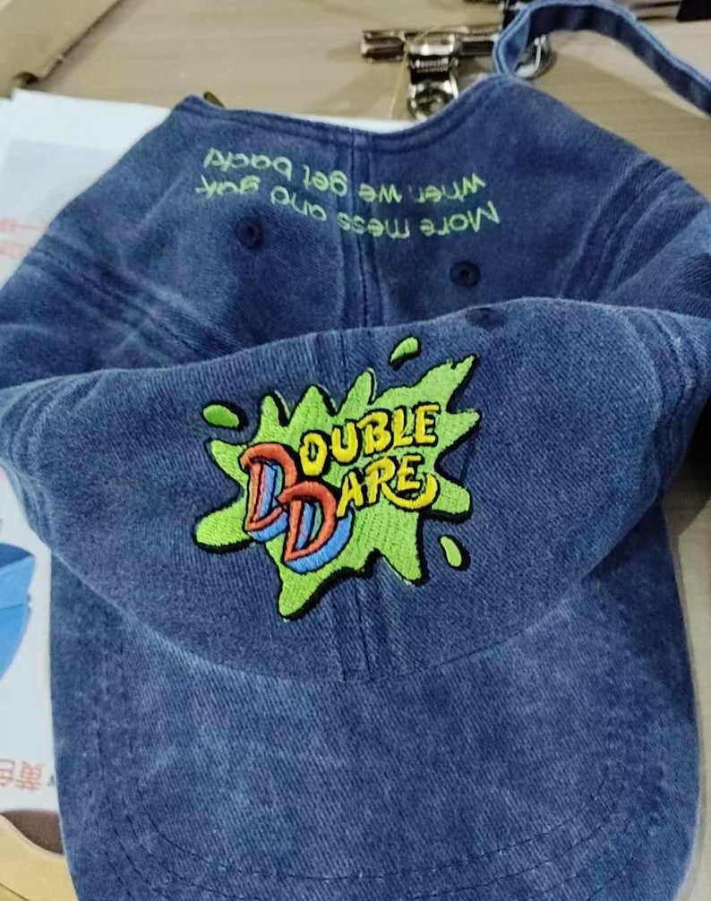 Puede incluir: Una gorra de b&eacute;isbol de mezclilla azul con un logotipo bordado en verde, amarillo y rojo que dice "Double Dare". La parte posterior de la gorra tiene texto blanco que dice "M&aacute;s desorden y m&aacute;s diversi&oacute;n, cuando volvamos"
