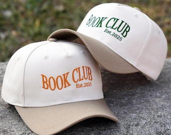 Pode incluir: Dois bonés de beisebol na cor branco off-white com abas bege. A parte frontal dos bonés apresenta texto bordado. Um boné tem texto laranja escrito BOOK CLUB Est.2025, enquanto o outro tem texto verde escuro escrito BOOK CLUB Est.2025.