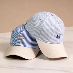 May include: Two baseball caps featuring a blue and white vertical striped pattern on the crown and a solid cream-coloured brim. One cap displays the embroidered initials NF in gold, while the other cap features the embroidered initials AM in blue.