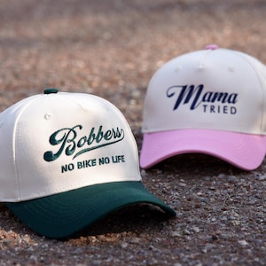 May include: Two white baseball caps with different coloured brims and embroidered text. The first cap has a green brim and says "Bobbers No Bike No Life". The second cap has a pink brim and says "Mama Tried".
