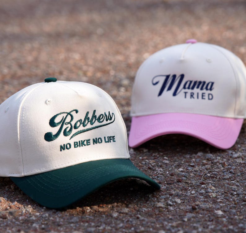 May include: Two white baseball caps with different coloured brims and embroidered text. The first cap has a green brim and says "Bobbers No Bike No Life". The second cap has a pink brim and says "Mama Tried".