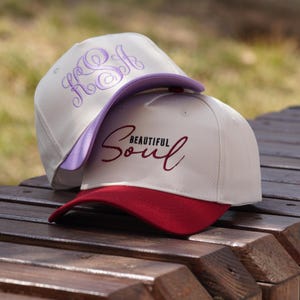 May include: Two white baseball caps with different embroidery. The top cap has a purple embroidered monogram. The bottom cap has a red embroidered phrase "Beautiful Soul".