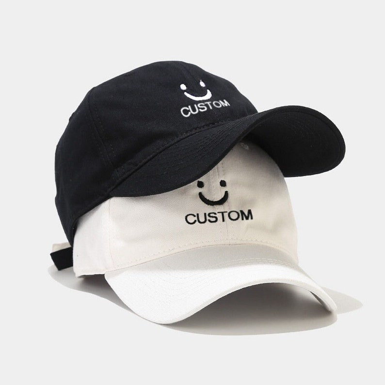 Puede incluir: Una gorra de b&eacute;isbol reversible en negro y blanco con una carita sonriente y la palabra "CUSTOM" bordada en ambos lados.
