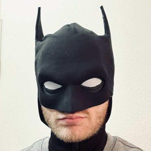 May include: A black Batman mask featuring white mesh eye coverings and distinctive pointed ears. The mask covers the upper face and head, with a cowl extending to the neck. The mask is crafted from a matte black fabric.