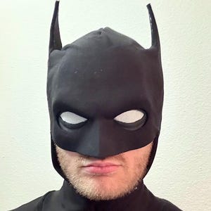 Op de afbeelding: Een zwart Batman-masker met witte mesh oogbedekkingen en puntige oren. Het masker bedekt het bovenste deel van het gezicht en het hoofd, met een donkere halslijn zichtbaar. Het masker is gemaakt van een mat materiaal.