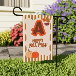 May include: A rectangular garden flag featuring vertical brown and white stripes. The centre displays a large brown letter A, the text HAPPY FALL Y'ALL in orange, and two pumpkins at the bottom. Three small autumn leaves are near the letter A.