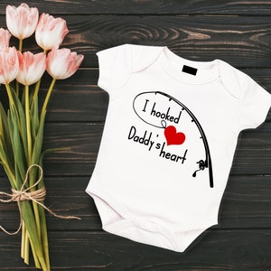 May include: White baby onesie with the text "I hooked Daddy's heart" in black script, a red heart, and a fishing rod graphic. A bouquet of pink tulips tied with twine sits to the left on a dark wood surface.