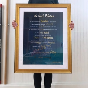 Peut inclure: Une impression encadr&eacute;e avec un fond bleu fonc&eacute; et des lettres dor&eacute;es. Le texte dit "We teach Pilates" et comprend une citation sur l'aide aux gens &agrave; ressentir la libert&eacute; dans leur corps. La citation mentionne &eacute;galement l'entra&icirc;nement des corps qui sont vuln&eacute;rables et tout le monde entre les deux. La citation se termine par "Nous savons que le changement se produit par petites touches au fil du temps et qu'am&eacute;liorer le bien-&ecirc;tre d'une personne peut avoir un impact positif sur toute la communaut&eacute;."