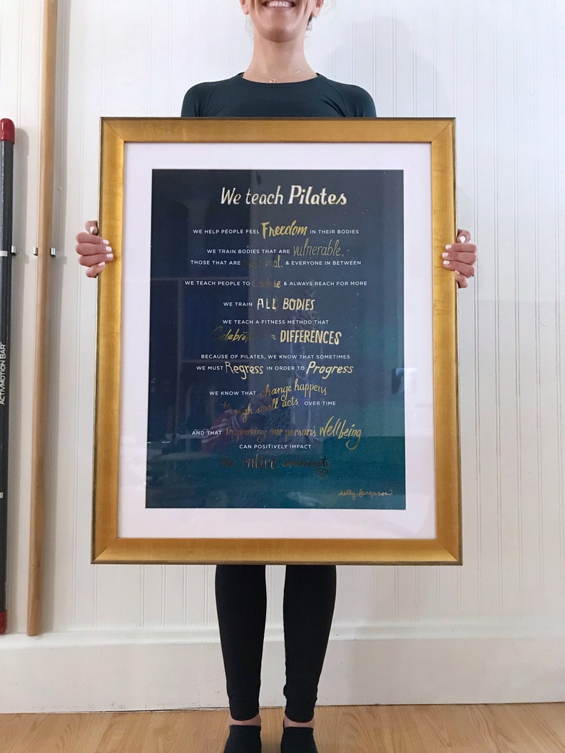 Peut inclure: Une impression encadr&eacute;e avec un fond bleu fonc&eacute; et des lettres dor&eacute;es. Le texte dit "We teach Pilates" et comprend une citation sur l'aide aux gens &agrave; ressentir la libert&eacute; dans leur corps. La citation mentionne &eacute;galement l'entra&icirc;nement des corps qui sont vuln&eacute;rables et tout le monde entre les deux. La citation se termine par "Nous savons que le changement se produit par petites touches au fil du temps et qu'am&eacute;liorer le bien-&ecirc;tre d'une personne peut avoir un impact positif sur toute la communaut&eacute;."