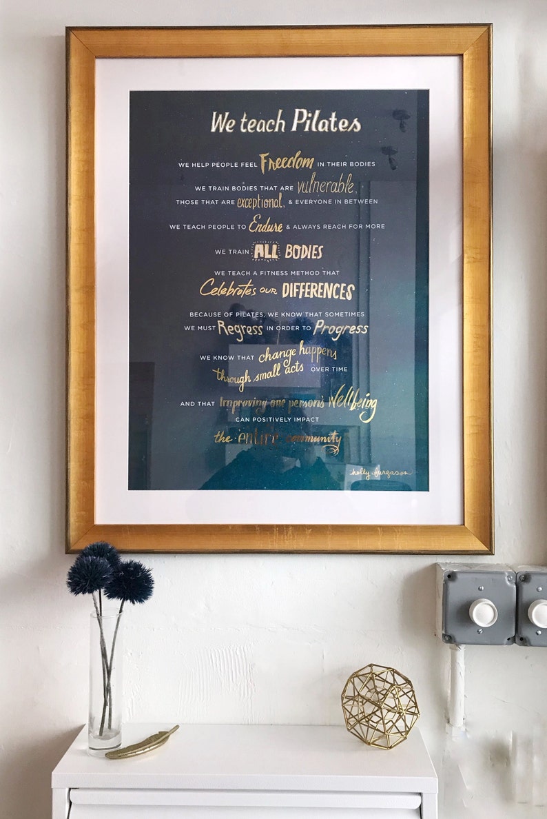 Peut inclure: Une affiche encadr&eacute;e avec un fond bleu fonc&eacute; et des lettres dor&eacute;es. L'affiche indique "We teach Pilates" et comprend une liste de valeurs et de croyances sur la pratique. Le texte comprend les mots "Freedom", "Vulnerable", "Exceptional", "Endure", "All Bodies", "Celebrates our Differences", "Regress", "Progress", "change happens", "Improving one person's Wellbeing", et "Pilates community".