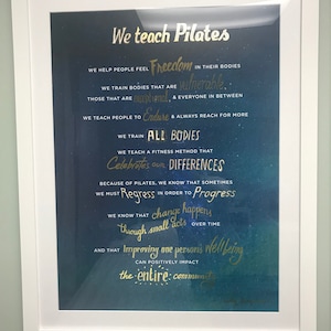 Peut inclure: Une affiche de motivation encadr&eacute;e avec un fond bleu et du texte dor&eacute;. Le texte dit "We teach Pilates" et comprend une citation sur l'aide aux gens &agrave; se sentir libres dans leur corps et &agrave; c&eacute;l&eacute;brer les diff&eacute;rences.