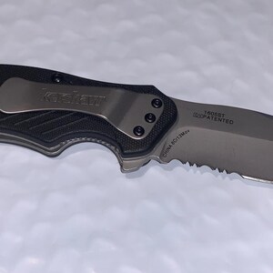 May include: A black and silver folding knife with a partially serrated blade. The knife has a textured black handle and a silver pocket clip. The blade has text that reads "TRIDENT" and "PATENTED".